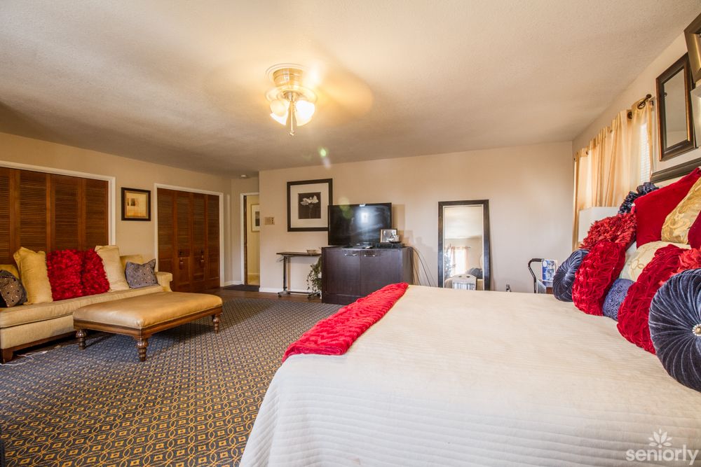 A spacious bedroom featuring a large bed with red accents, a flat-screen TV, and cozy furnishings.