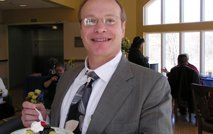 A man in a suit and glasses, smiling indoors.
