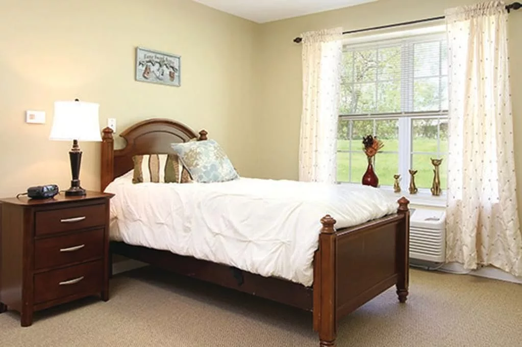A cozy bedroom featuring a wooden bed, a side table with a lamp, and a large window with sheer curtains.