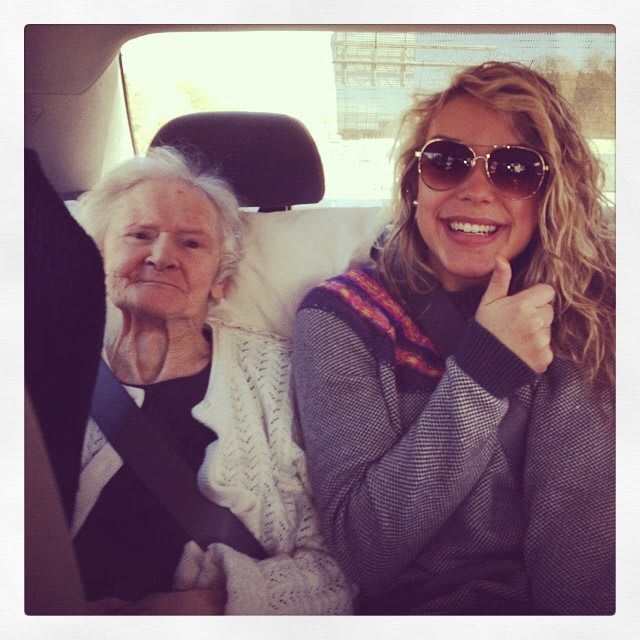 A young woman gives a thumbs up while sitting next to an elderly woman in the backseat of a car.