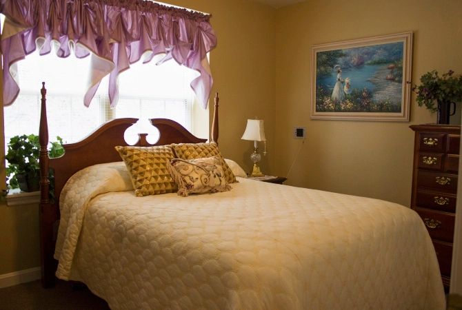 A cozy bedroom with a neatly made bed and decorative pillows, featuring a window with curtains and a bedside lamp.