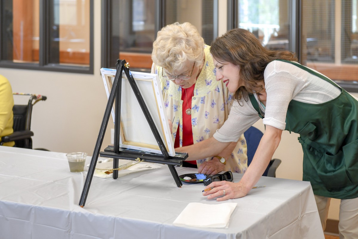 A younger woman assists an elderly woman in a painting class.
