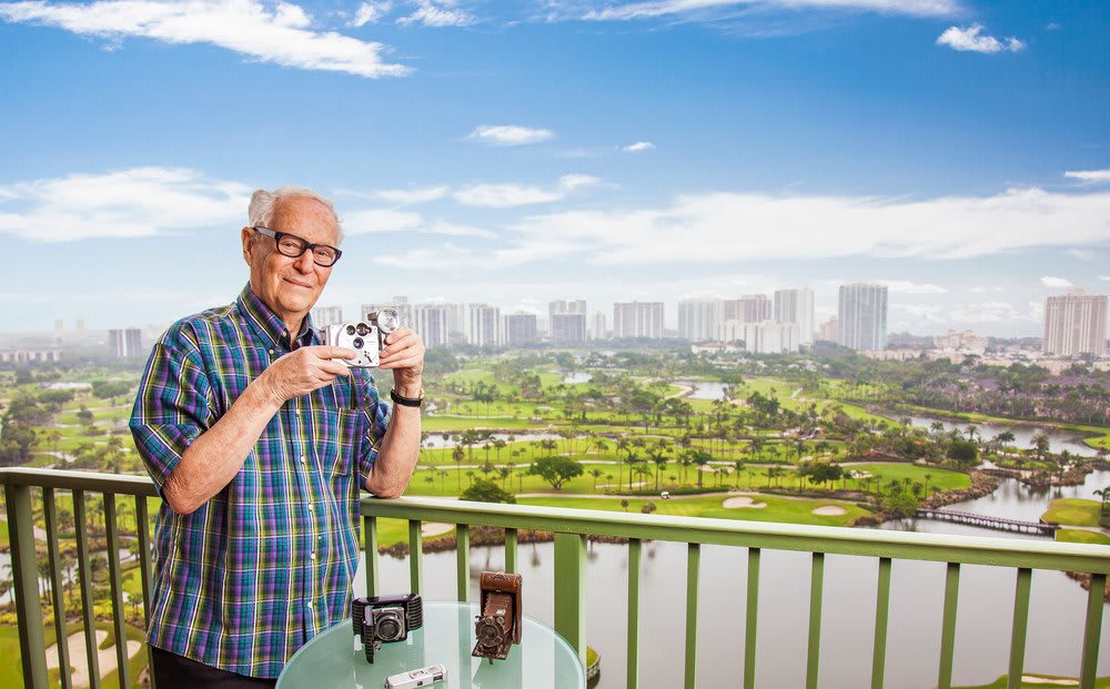 An elderly man stands on a balcony holding a camera, with a city skyline and lush greenery in the background.