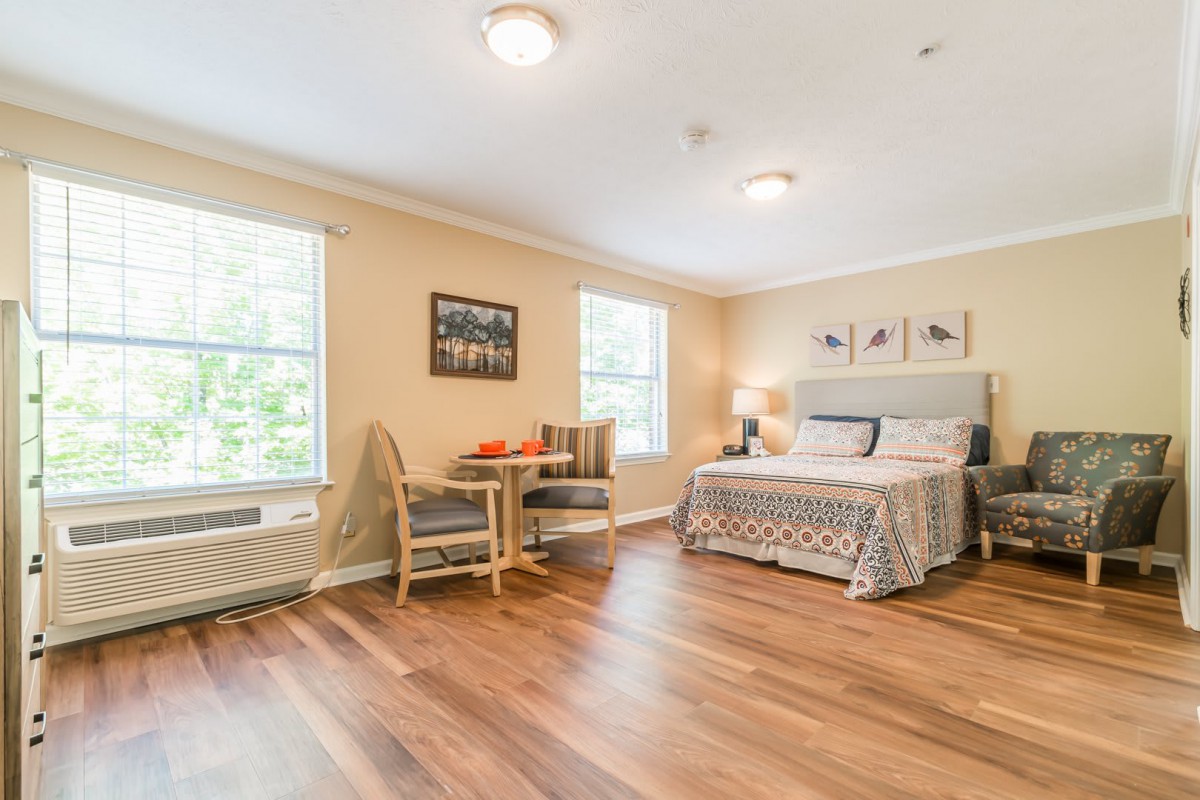 A spacious bedroom with hardwood floors, featuring a comfortable bed, a small table with chairs, and natural light from two windows.