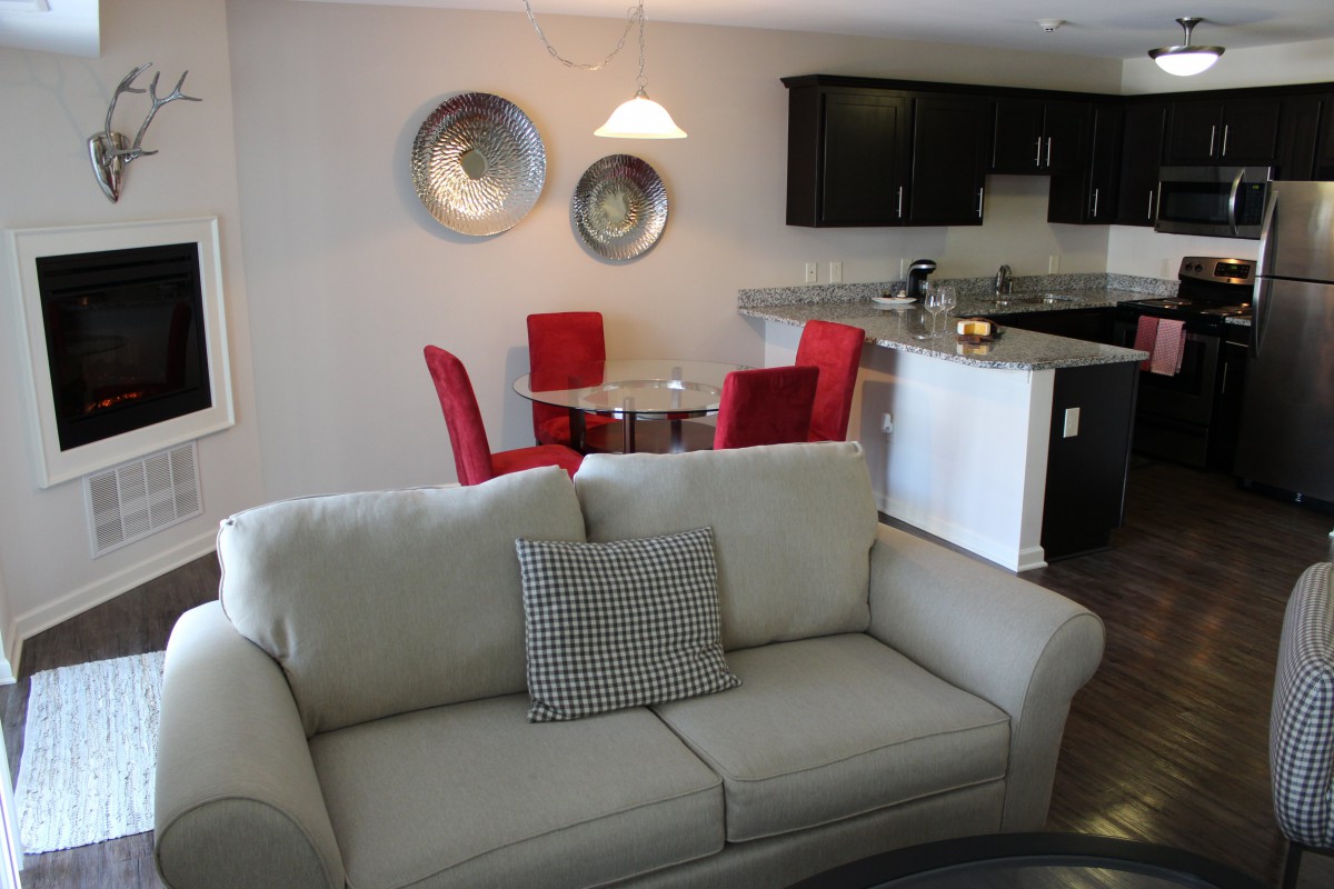 A modern living room with a sofa and cushion, adjacent to a kitchen area featuring red dining chairs.