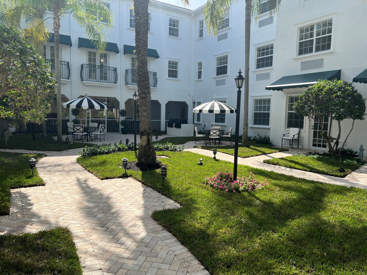 A charming courtyard with outdoor seating and palm trees, surrounded by a white building with green awnings.
