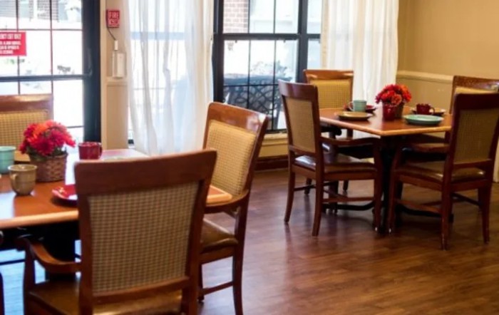 A cozy dining room with wooden tables and chairs, accented by bright natural light streaming through large windows with curtains.