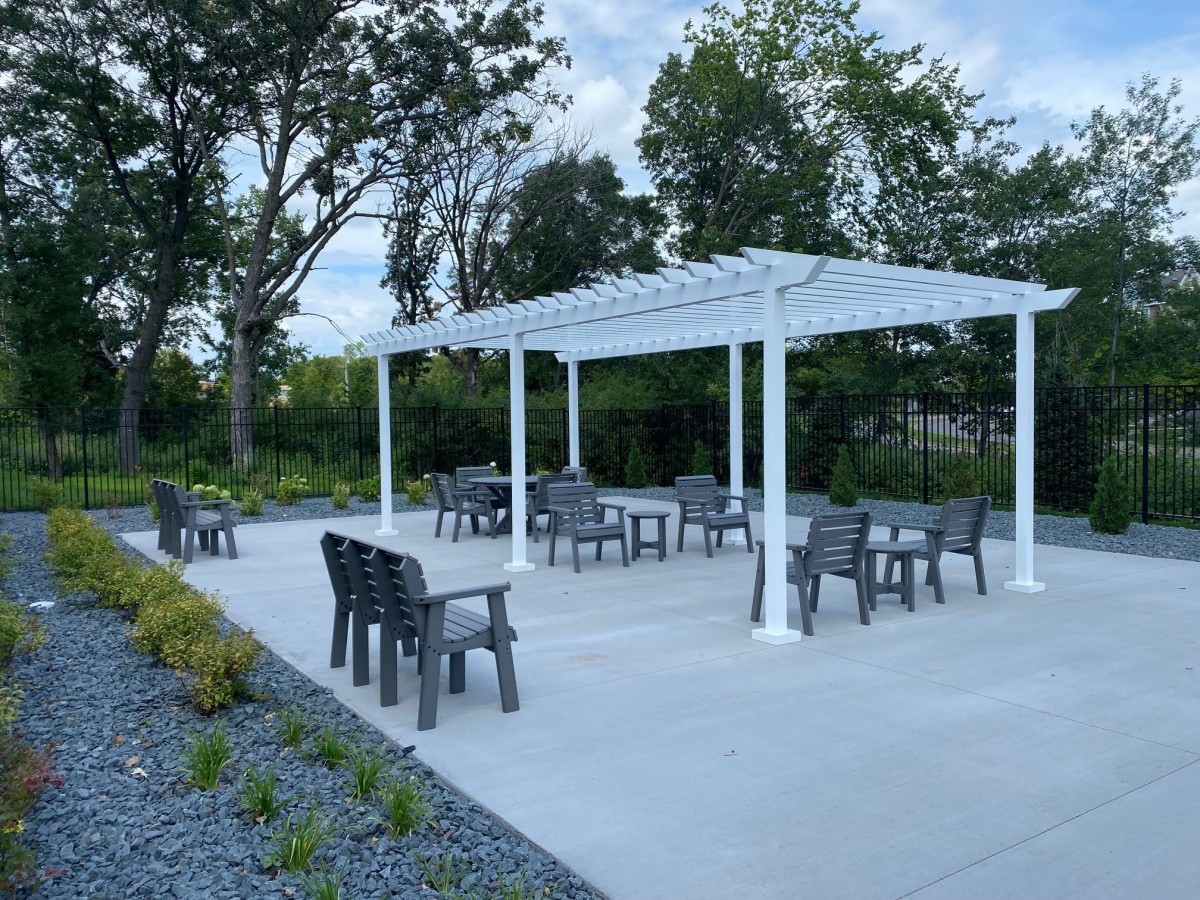An outdoor seating area with a white pergola and patio furniture, surrounded by trees and landscaping.