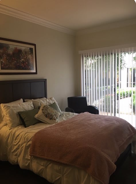 A cozy bedroom featuring a bed with pillows and a blanket, a large window with vertical blinds, and artwork on the wall.