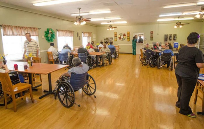 A community room in a nursing home with several seniors seated in wheelchairs.
