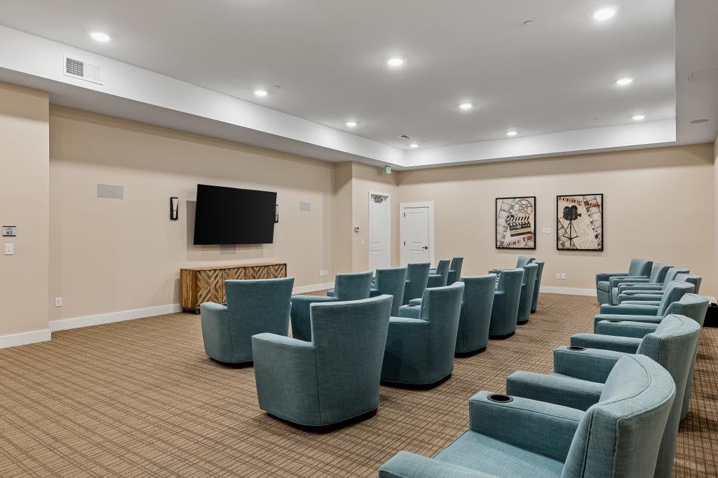 A modern home theater room with rows of comfortable chairs facing a large television, decorated with framed artwork and a carpeted floor.