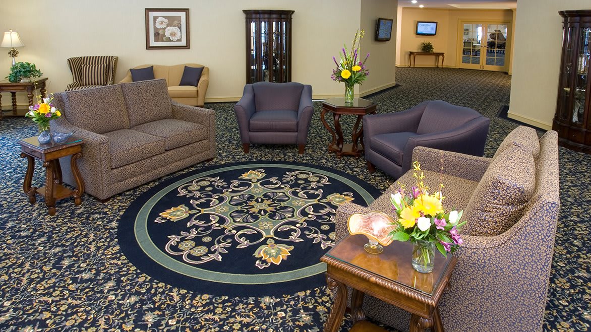 A cozy living room with sofas and armchairs arranged around a decorative rug, featuring a wooden table adorned with flower arrangements.
