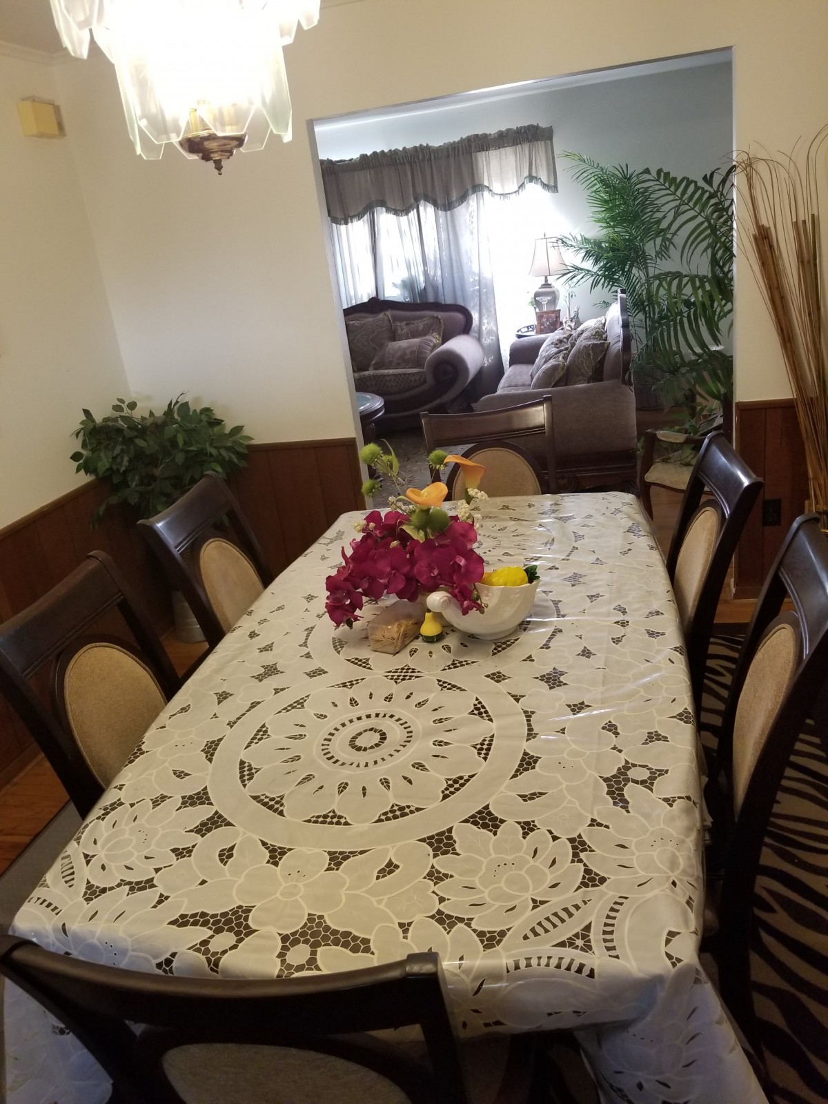 A dining room with a table covered in a decorative lace tablecloth, adorned with a floral centerpiece, and surrounded by cushioned chairs.