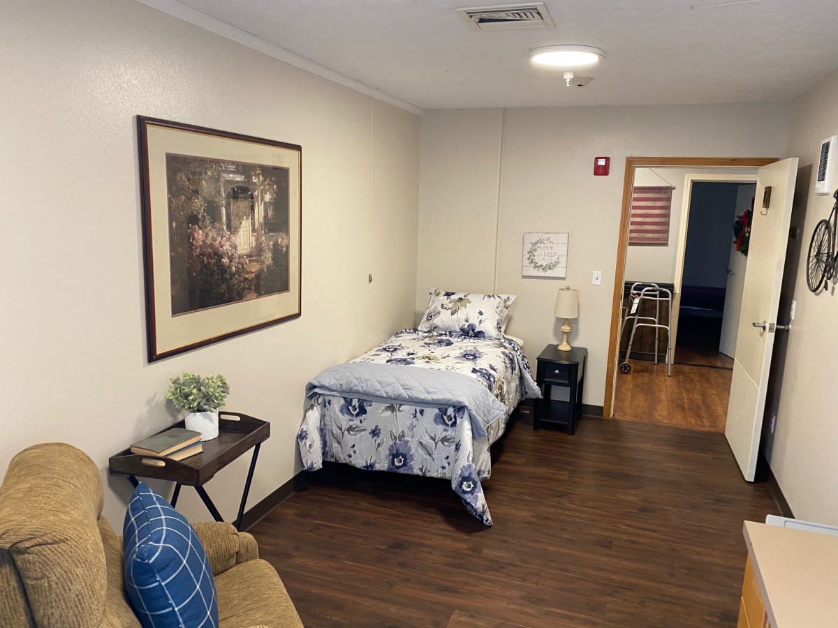 A cozy bedroom featuring a single bed with floral bedding, a side table with a lamp, and a comfortable chair. The room is decorated with a framed painting and has wooden flooring.