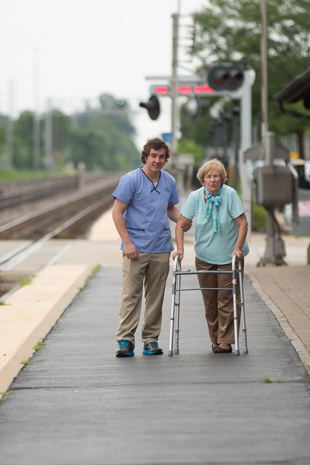 A caregiver assisting an elderly person with a walker at a train station.