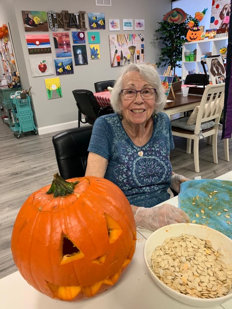 A woman smiles while carving a pumpkin indoors, surrounded by art supplies.