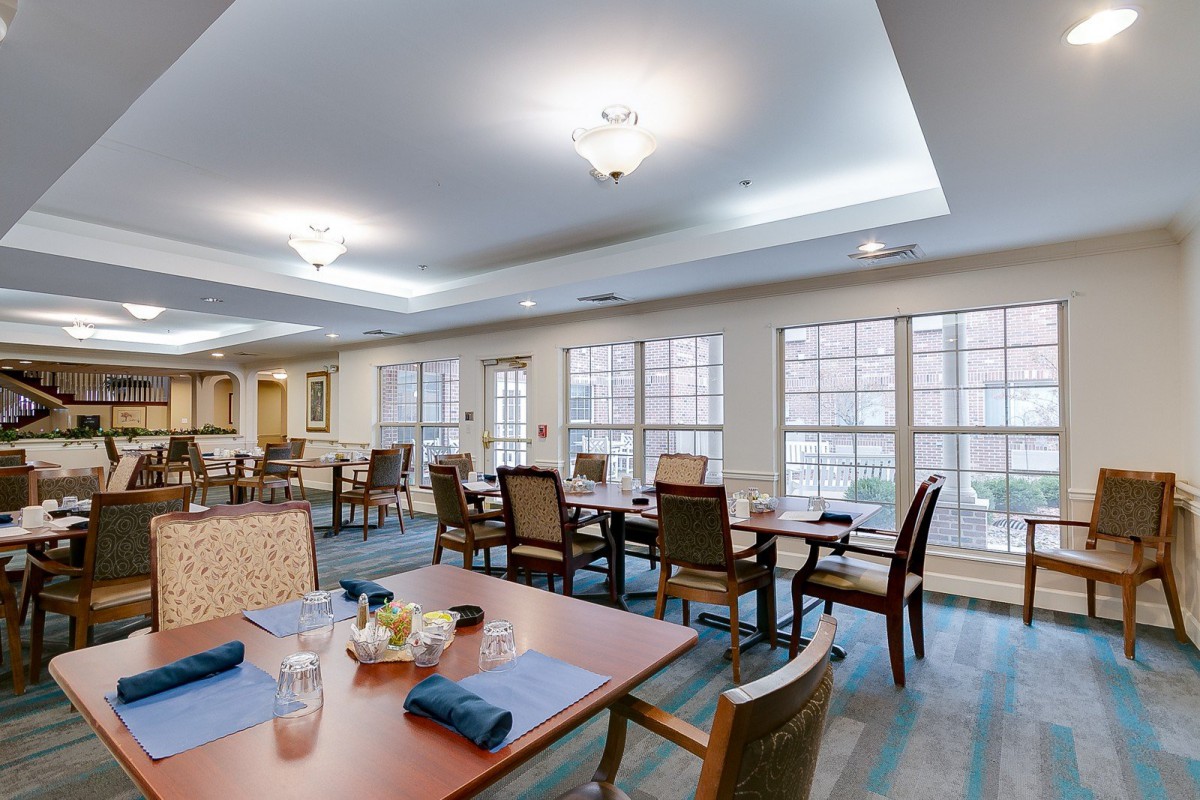 A spacious dining area with neatly arranged tables and chairs, illuminated by ceiling lights and natural light from large windows.