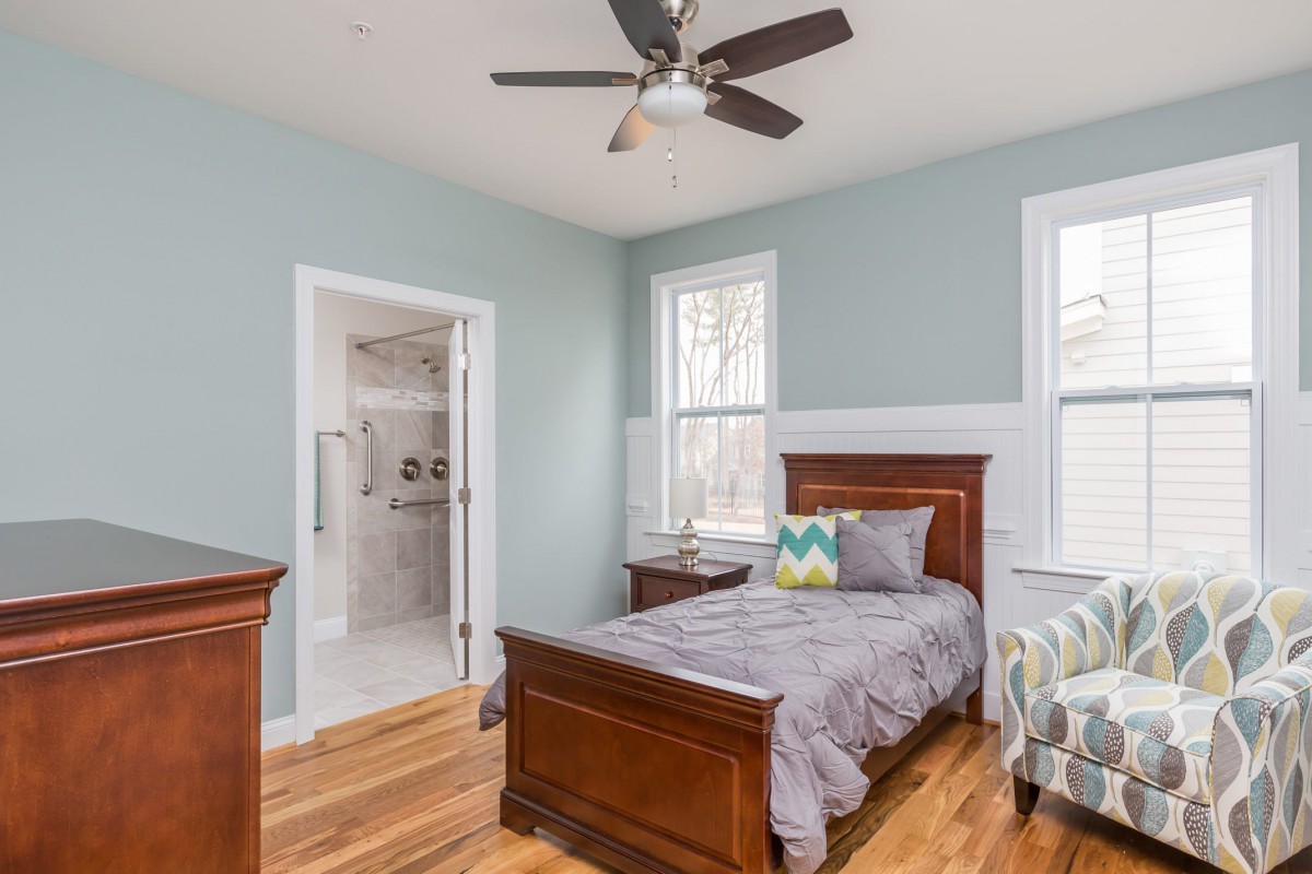 A cozy bedroom with a wooden bed and hardwood flooring, featuring a ceiling fan and adjacent bathroom.