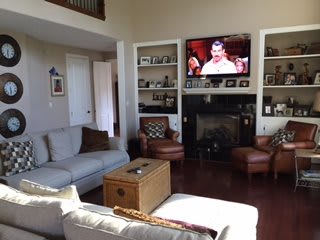 A cozy living room with a television above the fireplace, comfortable seating, and decorative shelving.