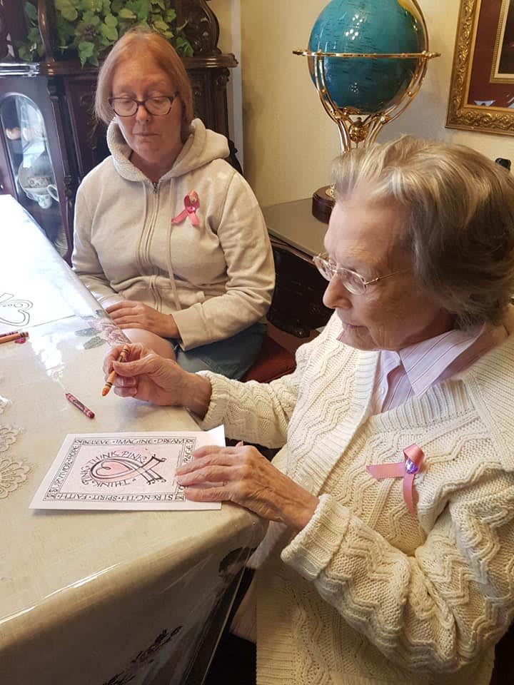 An elderly woman, along with another person, is engaged in crafting at a table indoors.