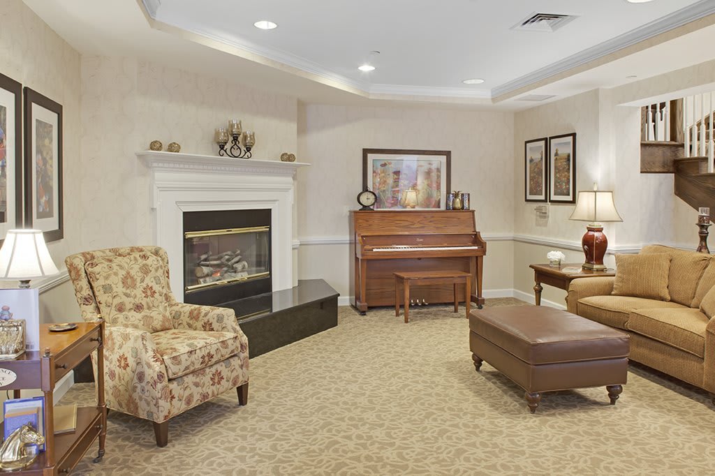 A cozy living room featuring a piano and a fireplace, complemented by an armchair and ottoman on a carpeted floor.