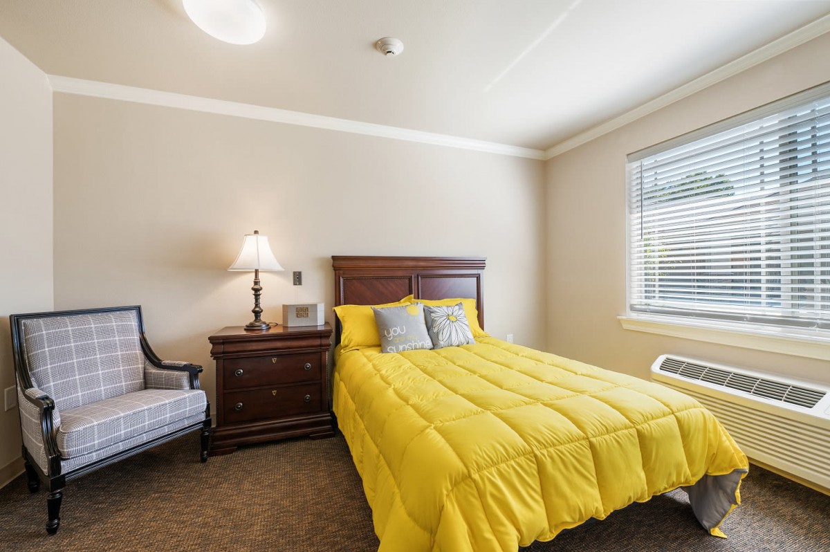 A cozy bedroom with a bright yellow comforter on the bed, accompanied by a nightstand with a lamp and an upholstered chair near the window.