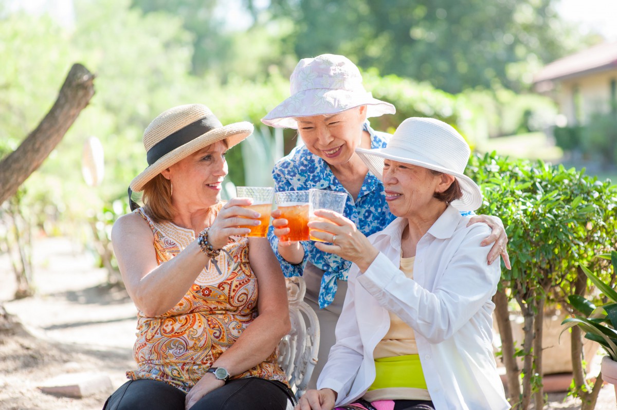 Three elderly women enjoying drinks together outdoors, wearing summer hats and smiling.