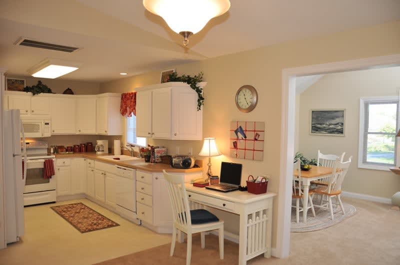 A cozy kitchen with a small home office setup, featuring white cabinets and a laptop on the desk.