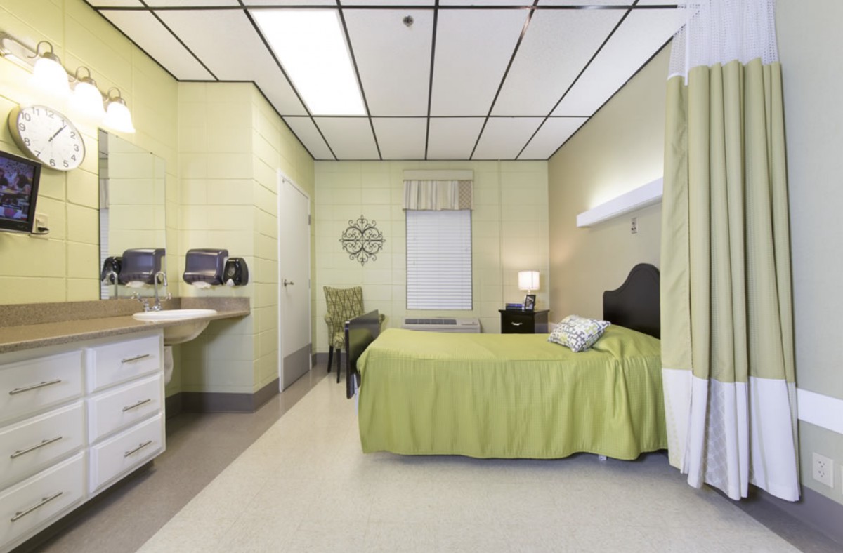 A modern hospital room featuring a single bed with green bedding, equipped with a sink and seating area.