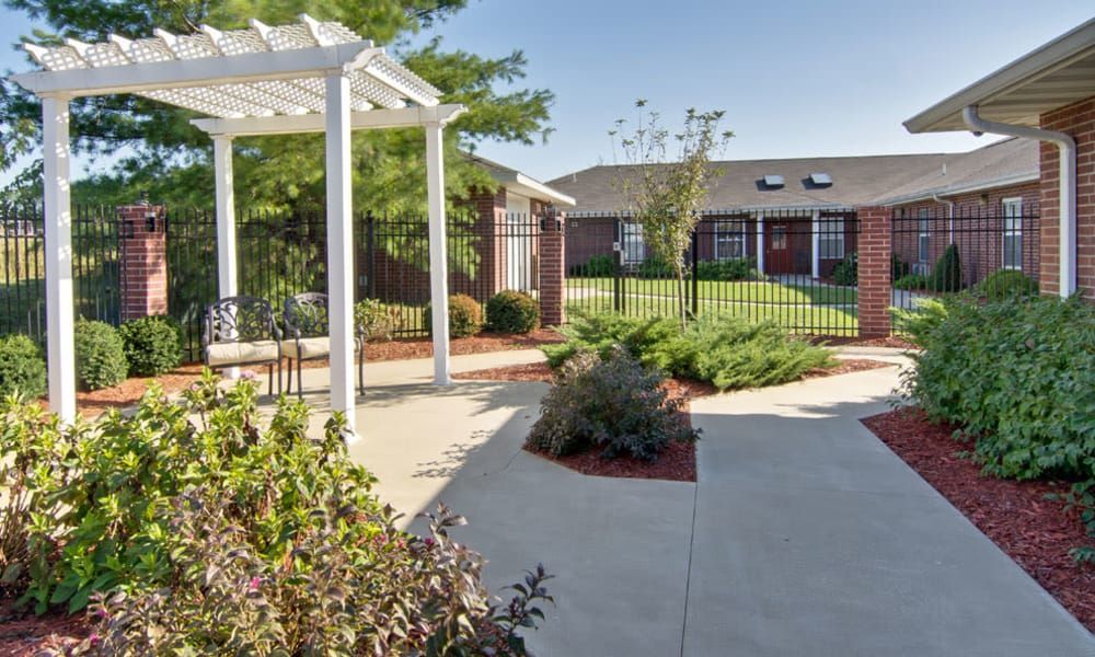 A landscaped garden with a pathway, pergola, and surrounding greenery leading to a brick building.
