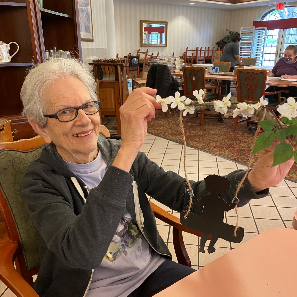 An elderly woman sitting indoors, engaging in a crafting activity with a decorated string.