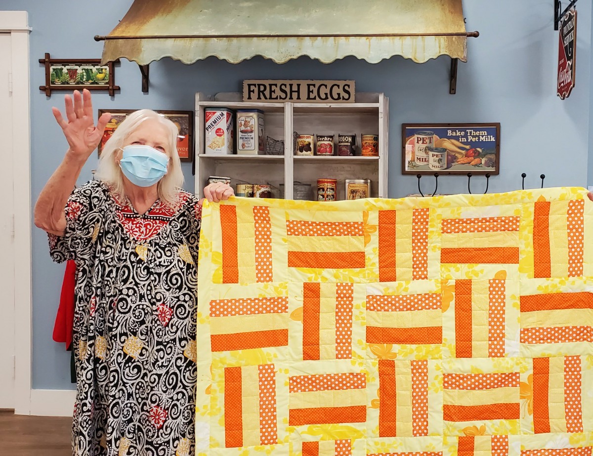 A woman wearing a mask proudly displays a handmade quilt with an orange and yellow pattern.