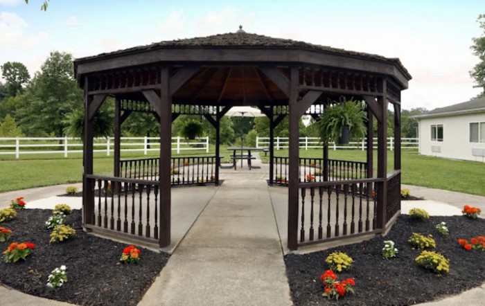 A wooden gazebo situated in a landscaped park with surrounding greenery and fences.