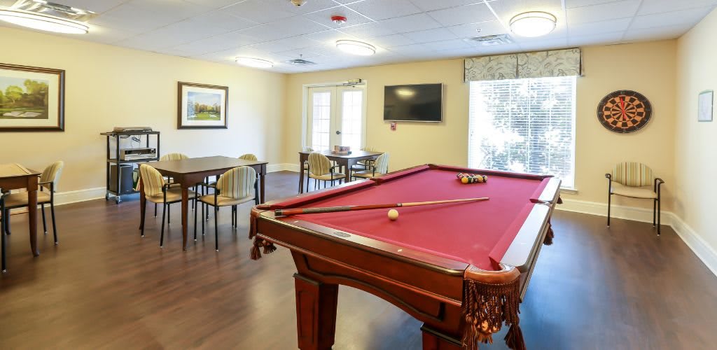A well-lit game room featuring a red-felt pool table, tables and chairs, and a wall-mounted television.