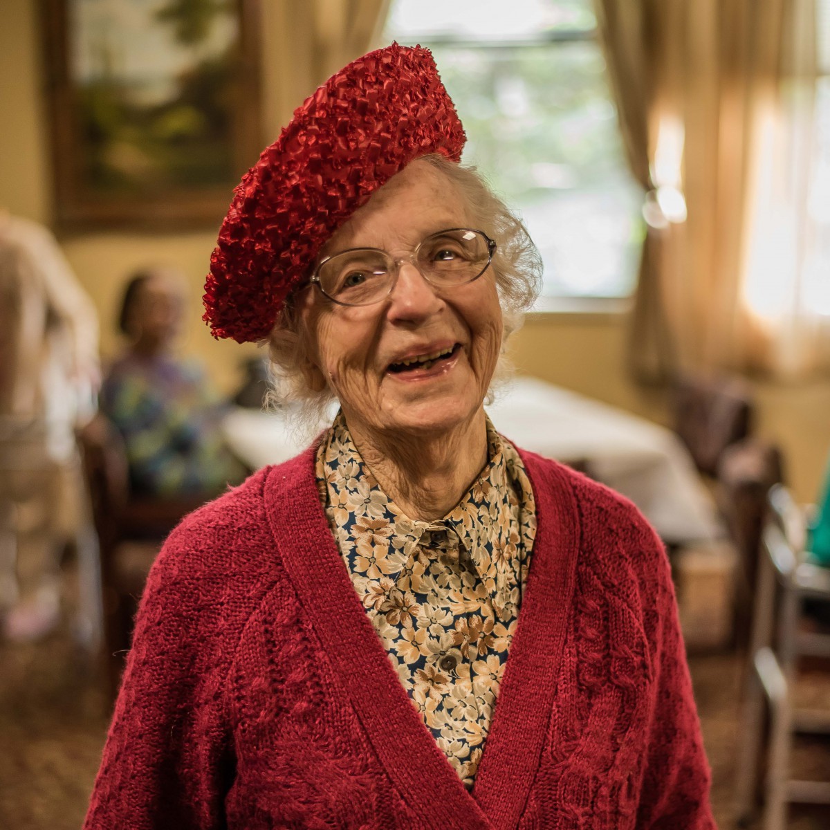 An elderly woman smiling warmly while wearing a red beret and sweater indoors.