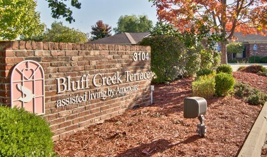 A brick wall sign for Bluff Creek Terrace, surrounded by trees with autumn leaves.