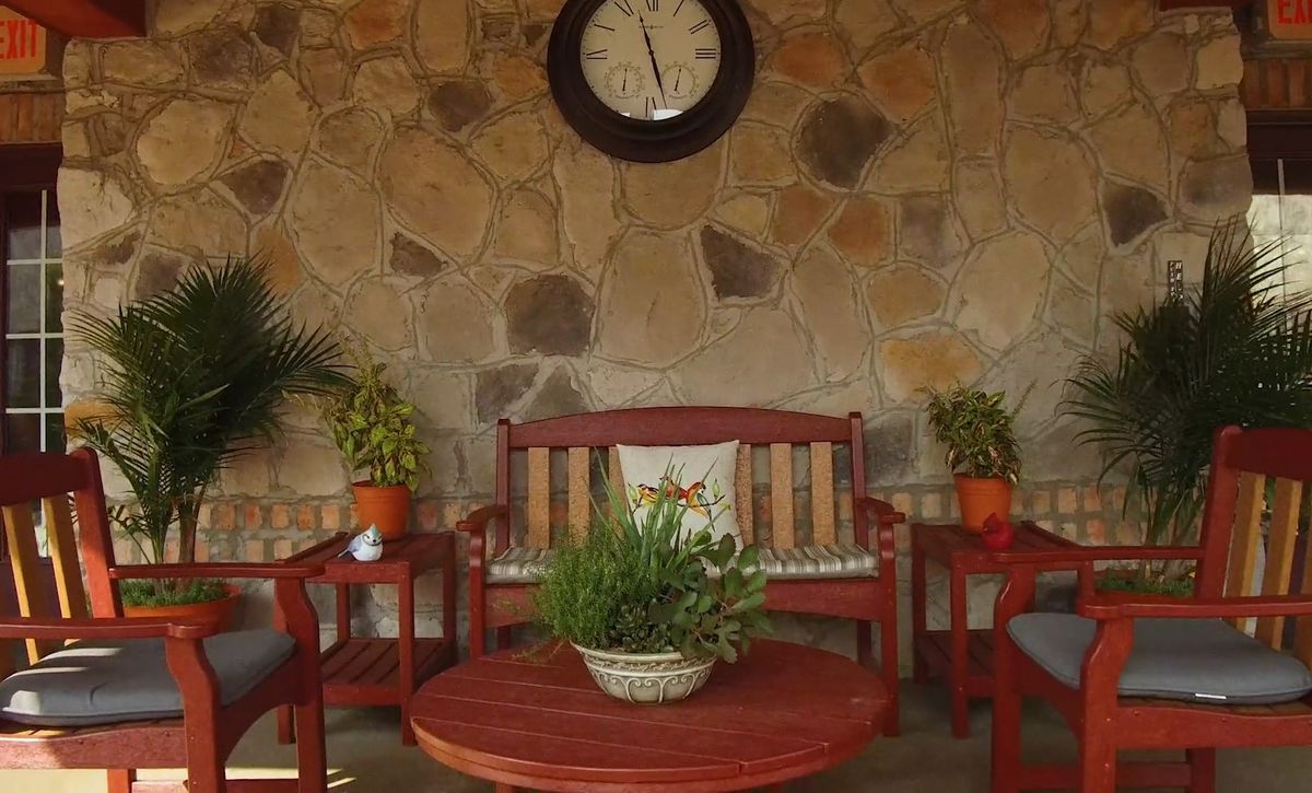 An inviting patio with wooden furniture, lush plants, and a decorative clock on a stone wall.