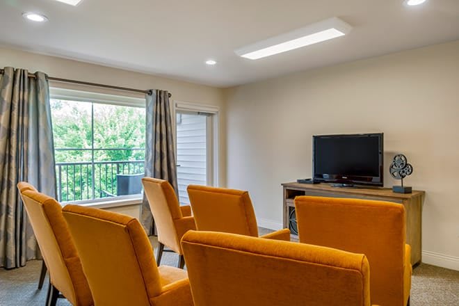A modern living room with mustard-colored chairs facing a television, featuring large windows and natural light.