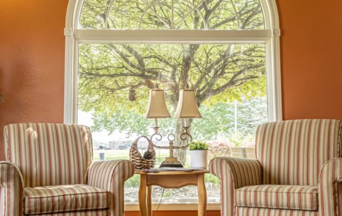 A cozy living room with two striped chairs, a small table with lamps, and a large window showcasing a view of a lush tree.