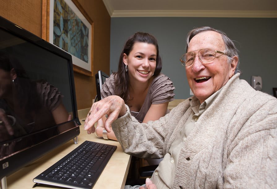 A young woman helping an elderly man with his computer, both smiling and enjoying the moment.