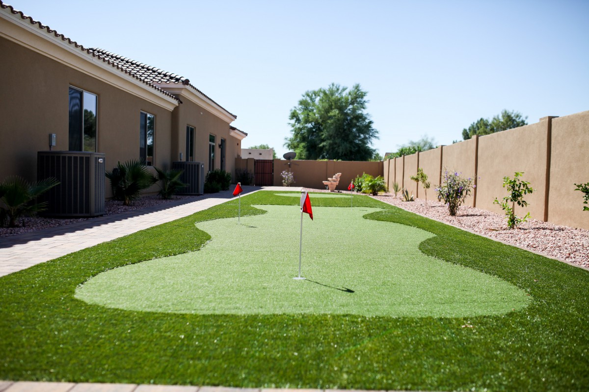 A backyard with an artificial grass putting green, featuring multiple flags and a pathway alongside a house.