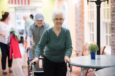 An elderly woman using a walker indoors, with a man following behind.