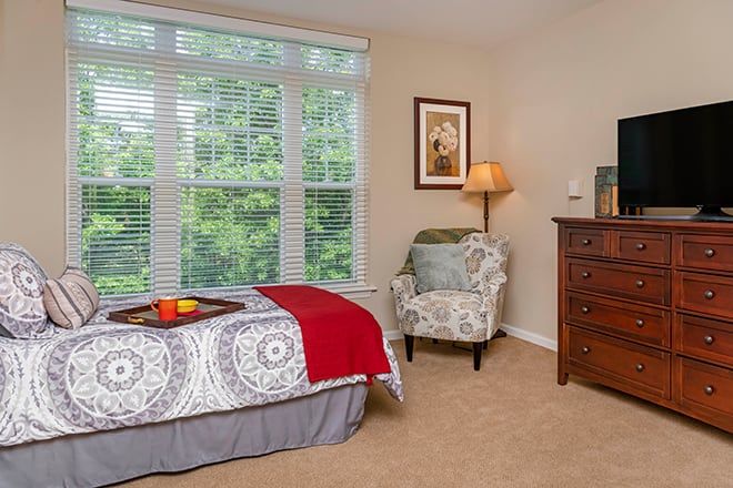 A cozy bedroom with a large window, featuring a bed with a patterned bedspread, an armchair, and a wooden dresser topped with a lamp.