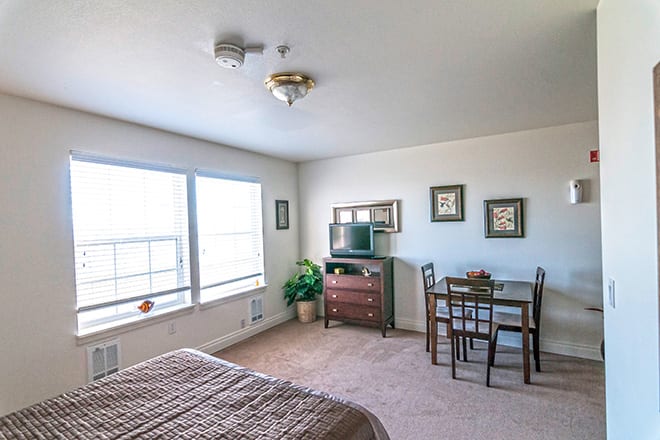 A bright and cozy bedroom with large windows, a bed, a small table with chairs, and a dresser equipped with a TV.