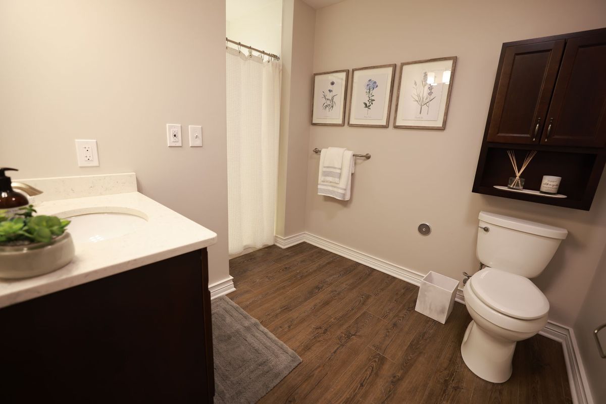 A modern bathroom with wooden flooring, featuring a sink, toilet, and shower area. Framed artwork decorates the wall above neatly hung towels.