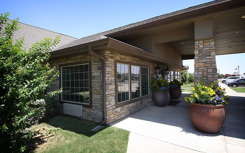 A modern house with a stone facade and large windows, surrounded by greenery on a sunny day.