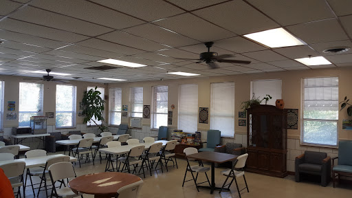 A spacious community room set up with tables and chairs, featuring a ceiling fan and natural light from the windows.
