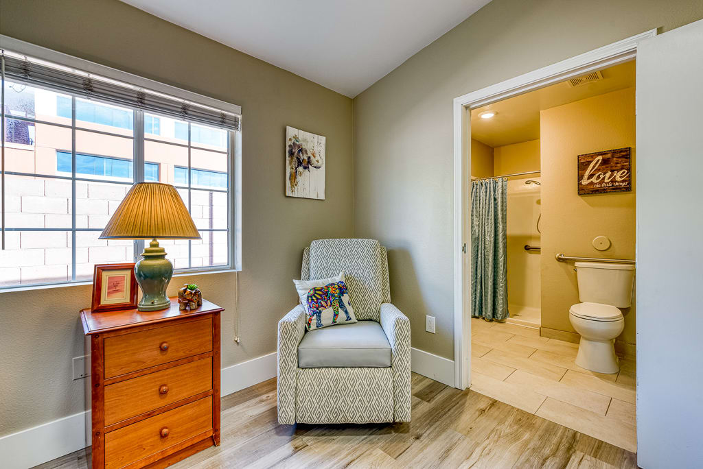 A cozy living space featuring an armchair, dresser with a lamp, and an adjoining bathroom.
