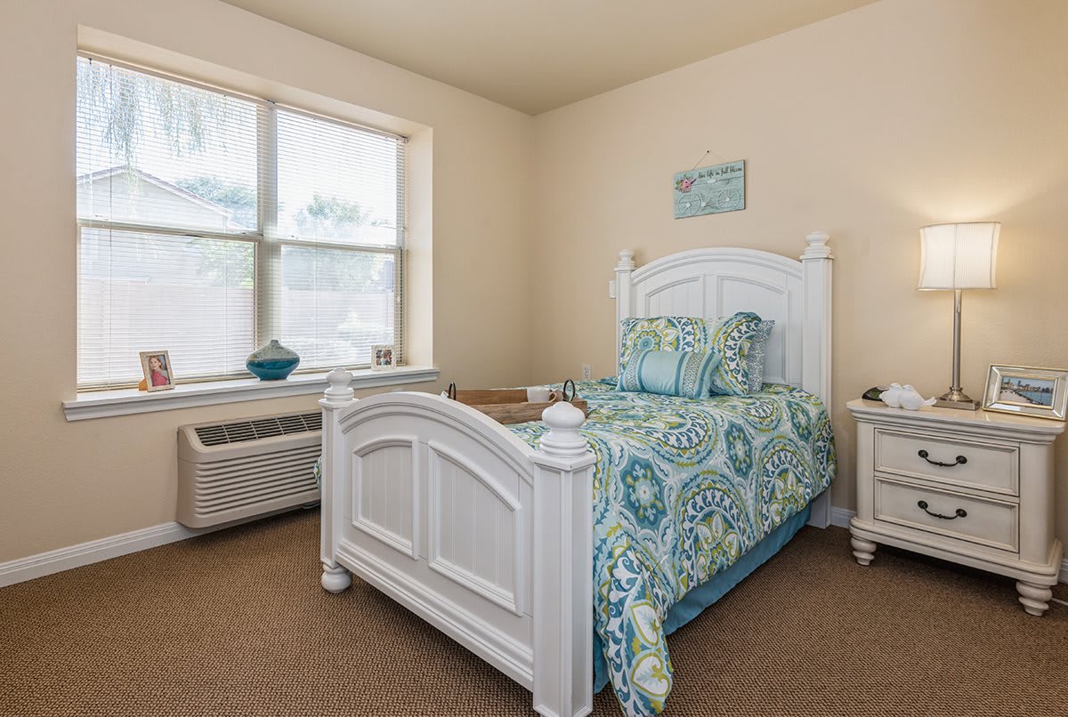 A cozy bedroom with a single bed and elegant white furniture, featuring patterned bedding and natural light from a window.