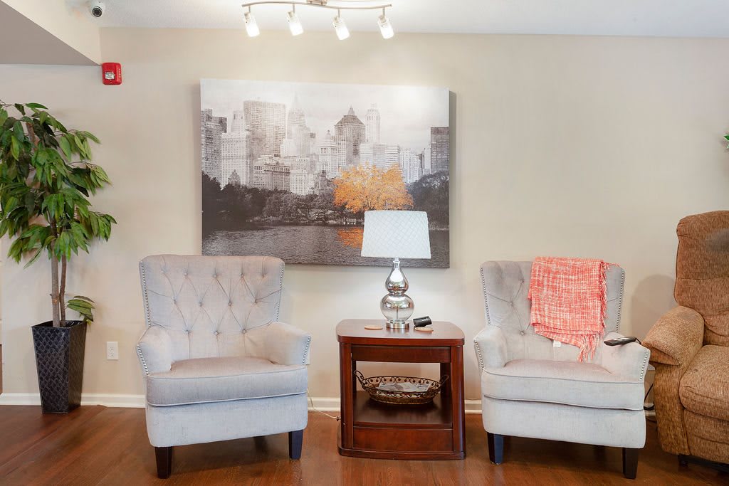 A cozy living room setup with two light grey chairs, a wooden side table with a lamp, and artwork on the wall.